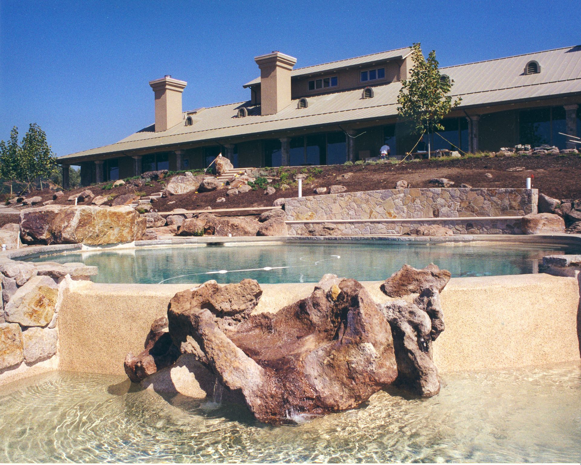 Rocky resort pool with clear water and a beige building in the background.
