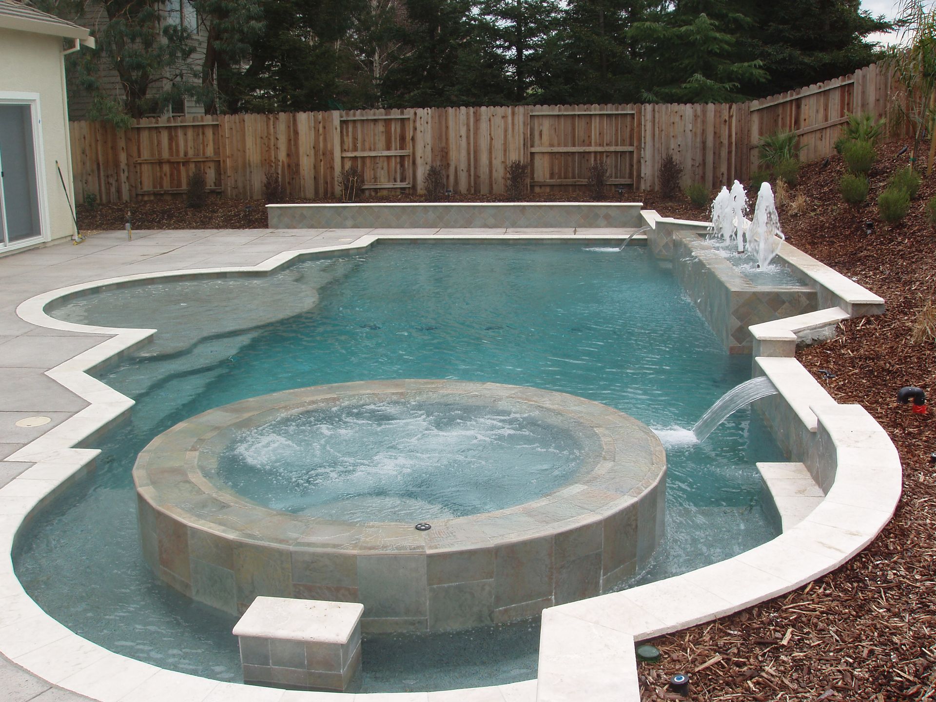 Backyard swimming pool with circular spa, turquoise water, white coping, and a wooden fence
