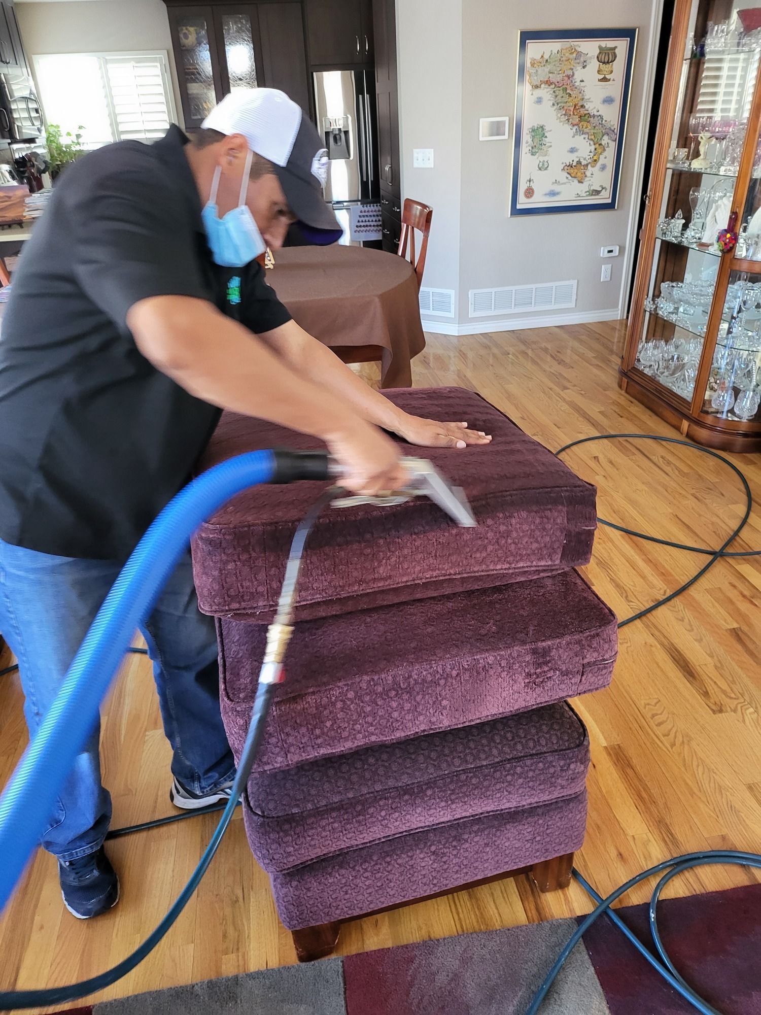 A man wearing a mask is cleaning a purple ottoman with a vacuum cleaner.