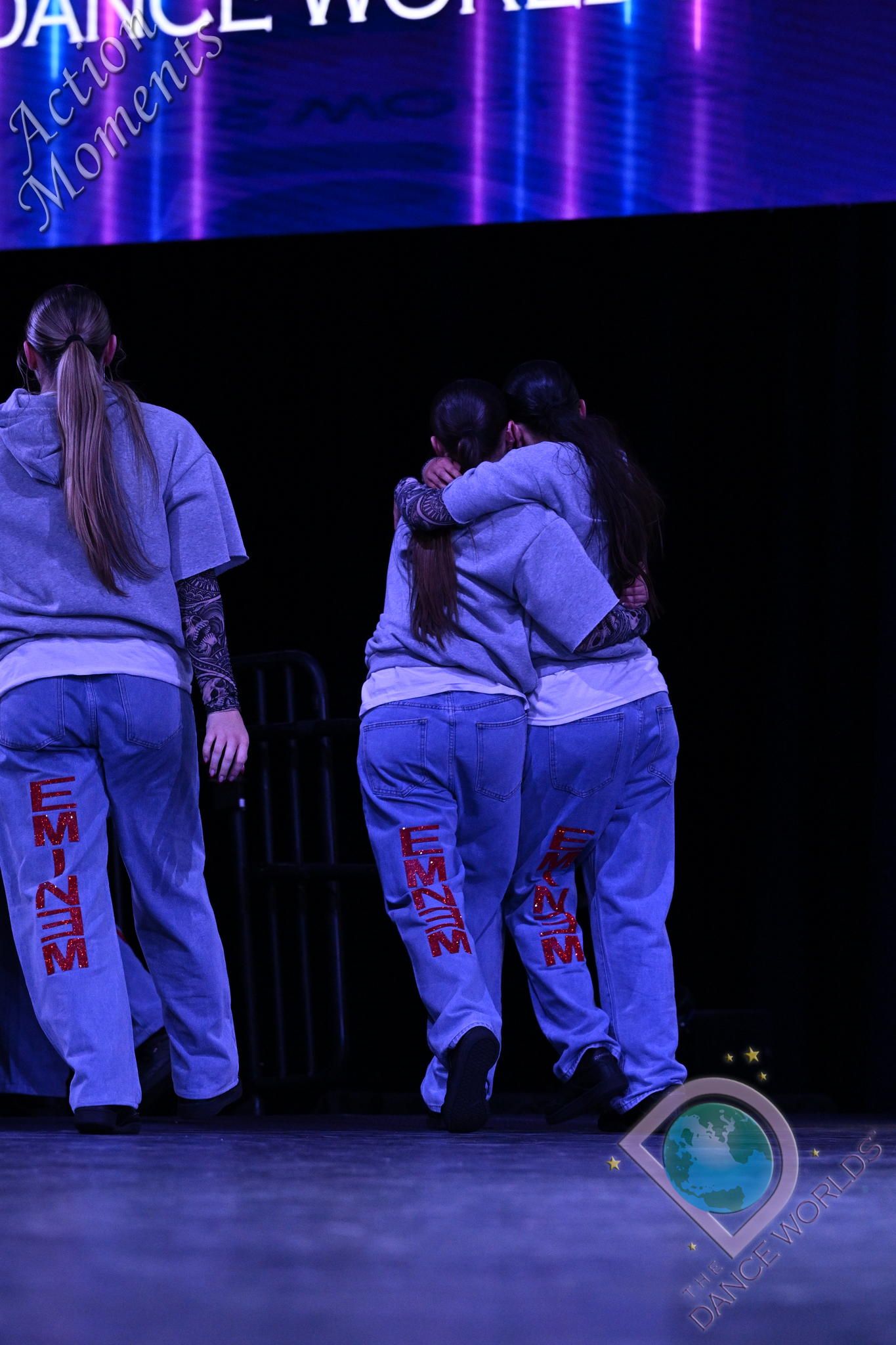 A group of women are hugging each other on a stage in front of a screen that says dance world