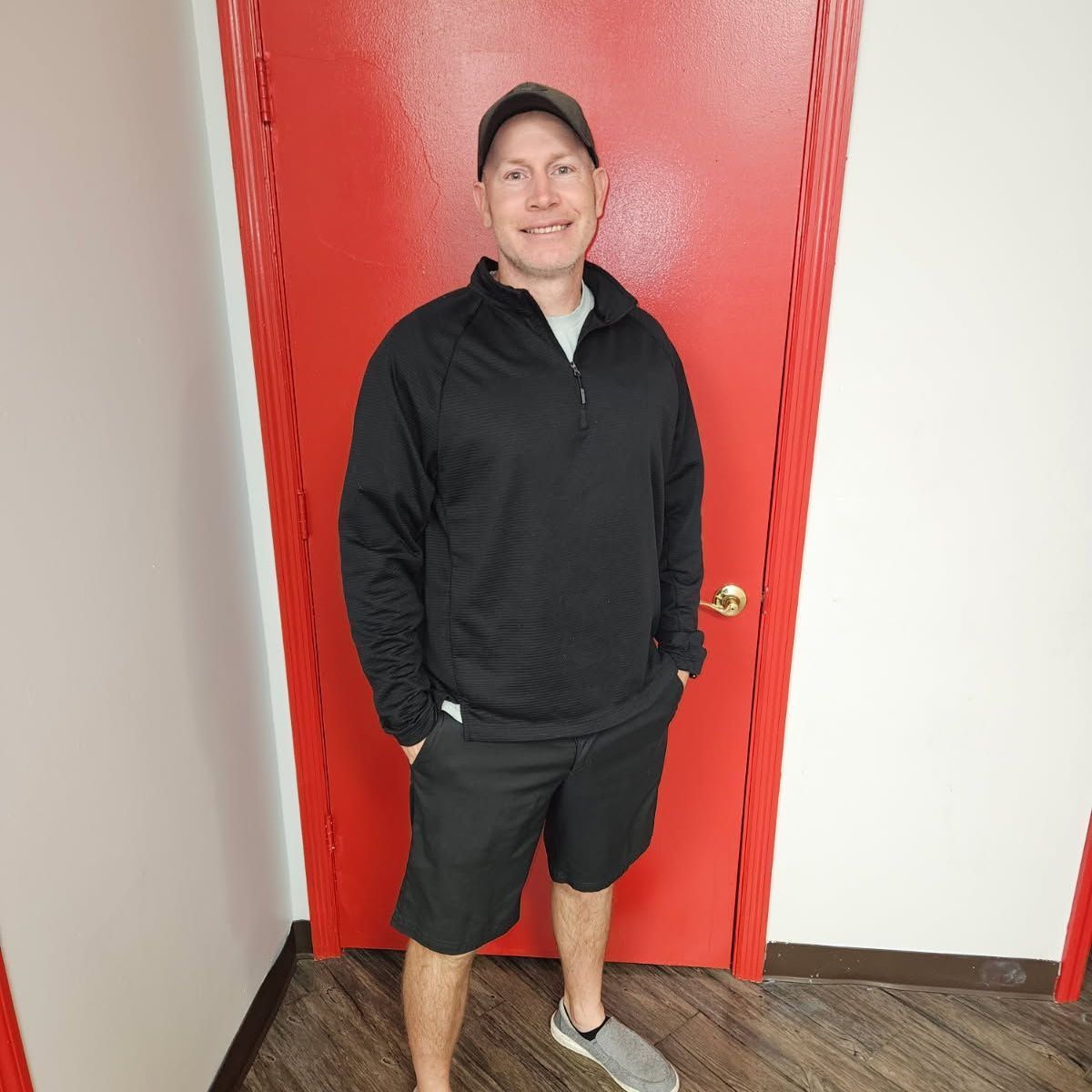 A man in a black shirt and shorts is standing in front of a red door