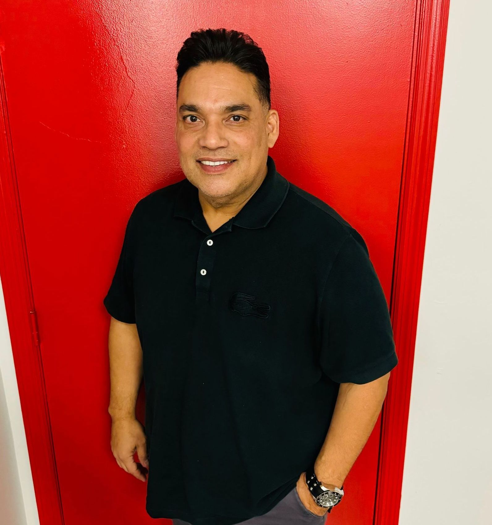 A man in a black shirt is standing in front of a red door.