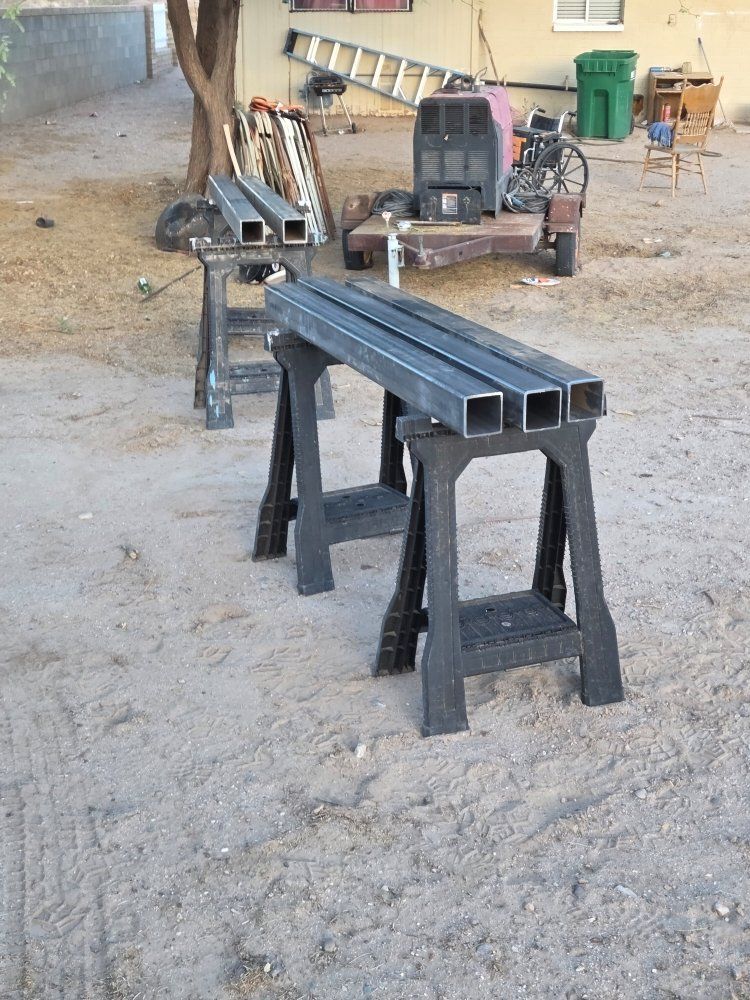 Square metal tubes on black sawhorses in a dirt yard, welding equipment and tools in the background.