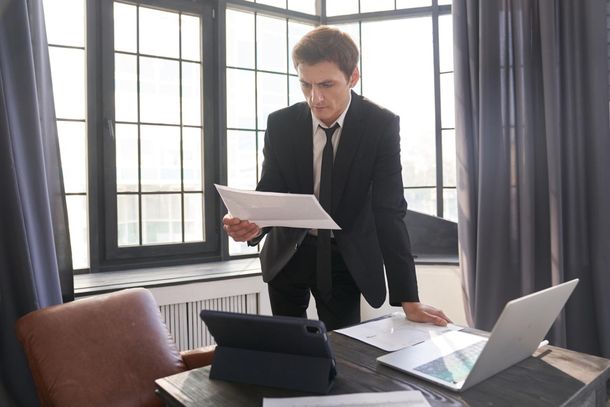Man in suit reviewing documents at a desk with a laptop and tablet, near a window.