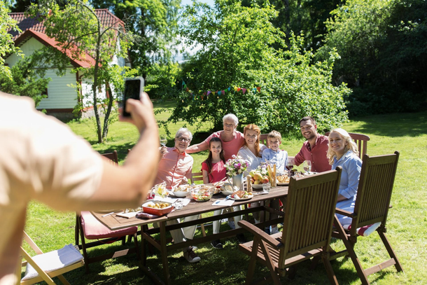 Person takes photo of family gathered around a table outdoors, set for a meal.