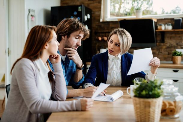 A woman in a blue blazer reviews paperwork with a couple at a kitchen table.