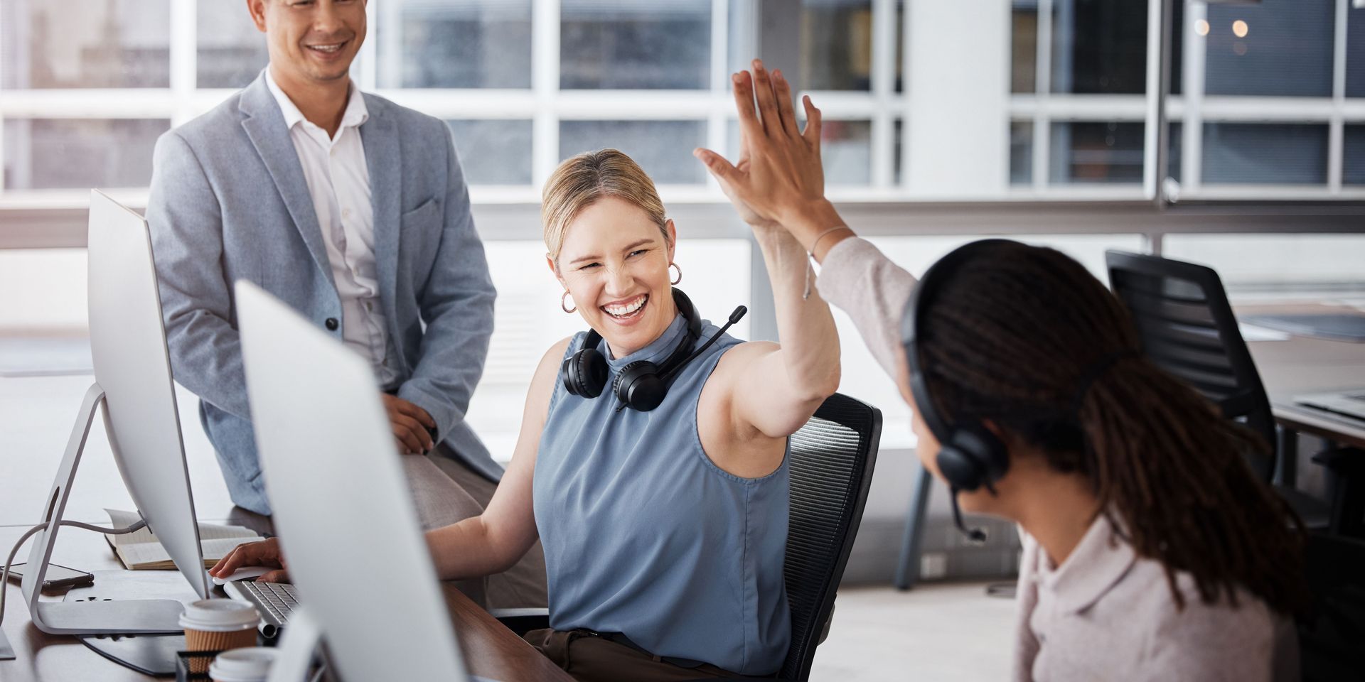 Three office workers celebrate success with a high five. Two women and a man are in an office setting.