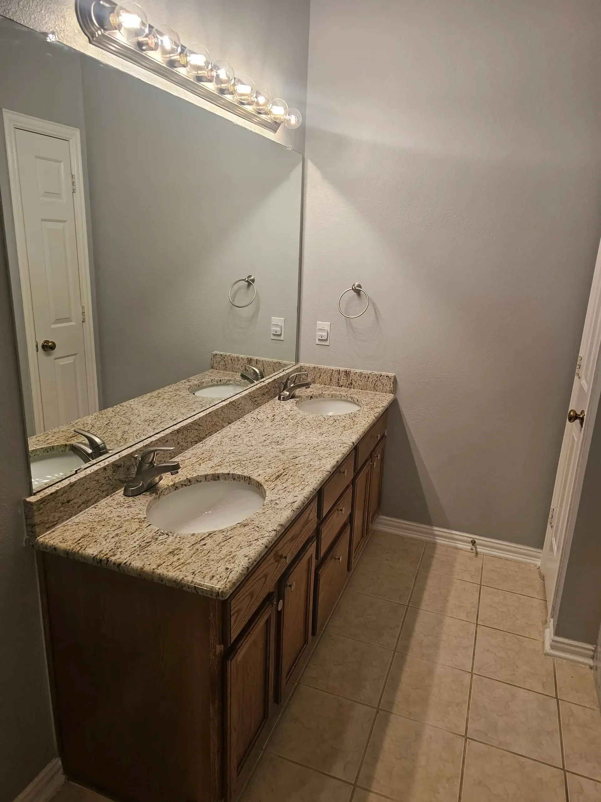 Bathroom with double sinks, light granite countertop, and tan cabinets, with beige tile flooring.