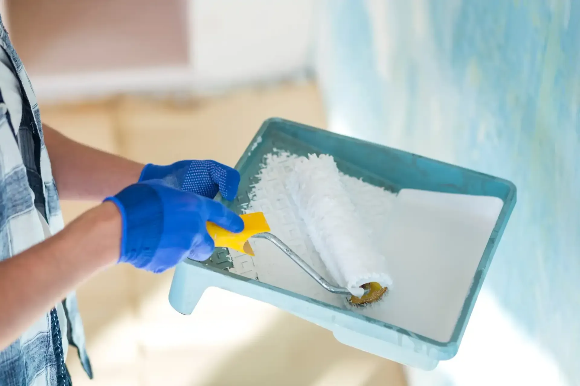 Person wearing blue gloves using a paint roller in a tray of white paint to paint a wall blue.