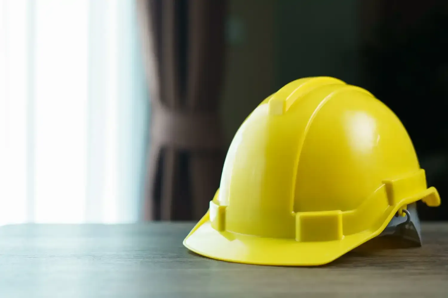 Yellow hard hat on a wooden surface, near a window with curtains.