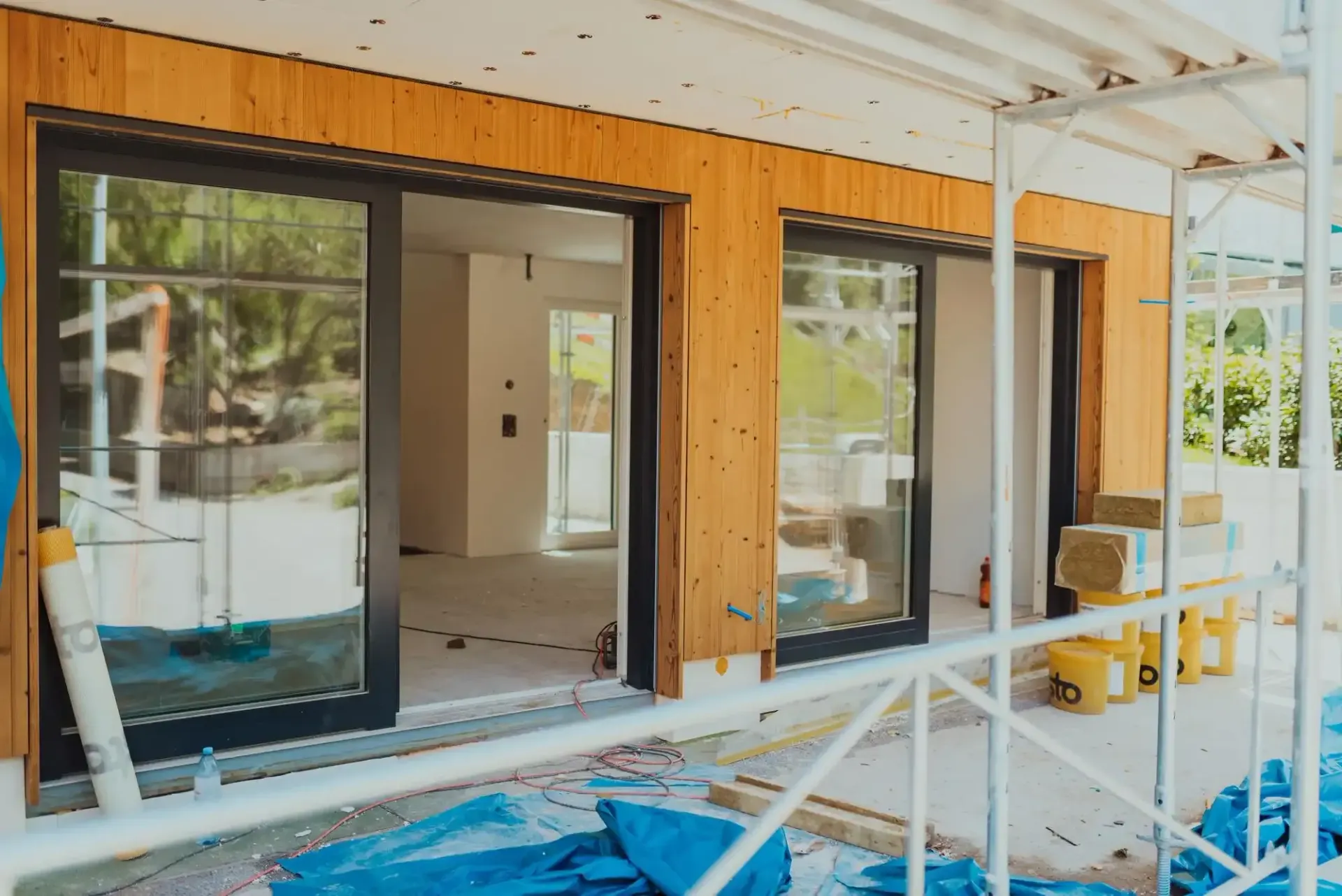 Exterior view of a building under construction, featuring sliding glass doors framed by wood panels, and scaffolding.