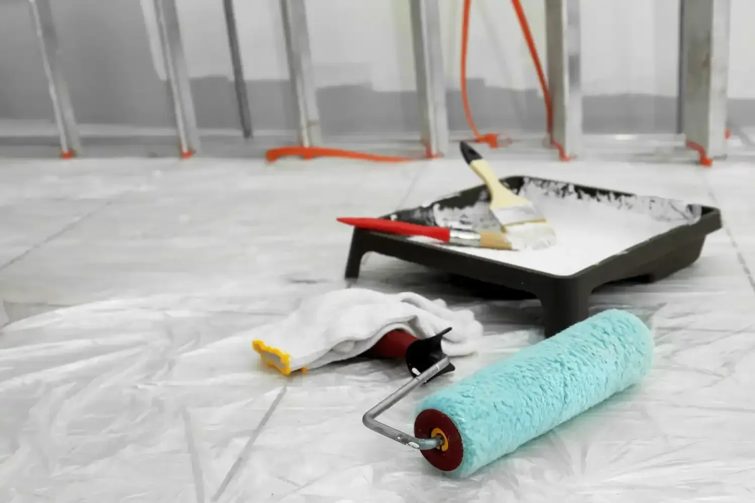 Paint tray with white paint, roller, brushes, work gloves, and ladder in room being painted.
