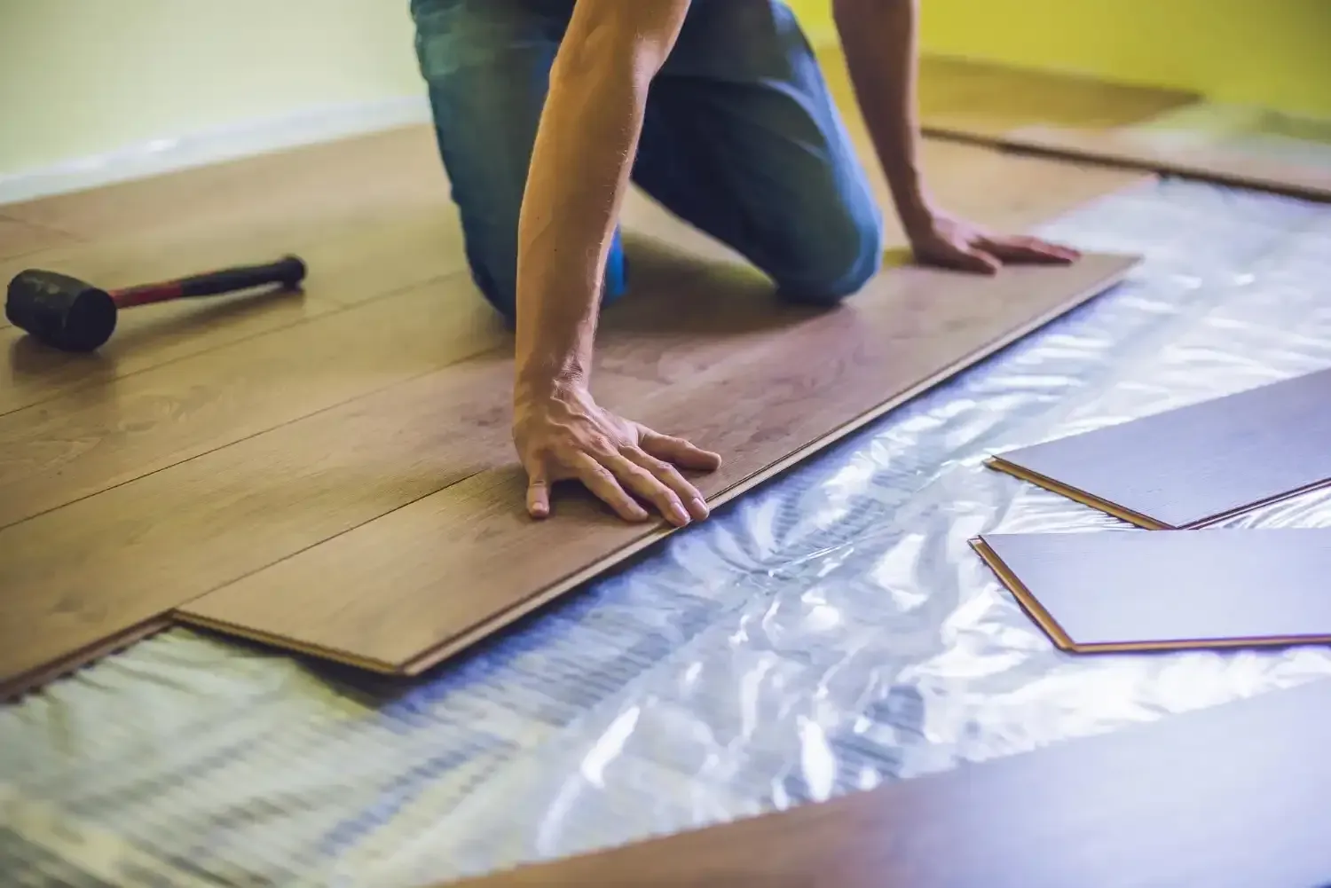 Person installing laminate flooring, kneeling, with tools and materials on subfloor.