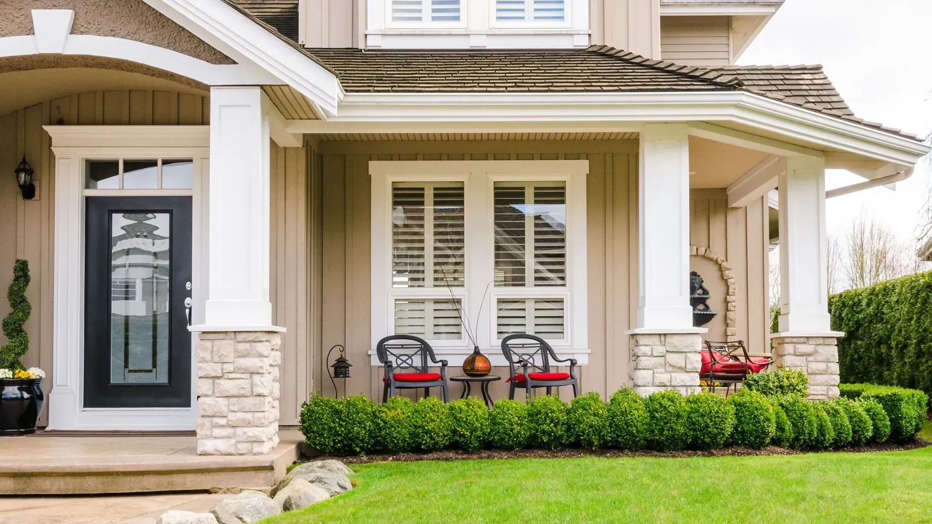 Beige house exterior with front porch, pillars, black door, and green lawn.