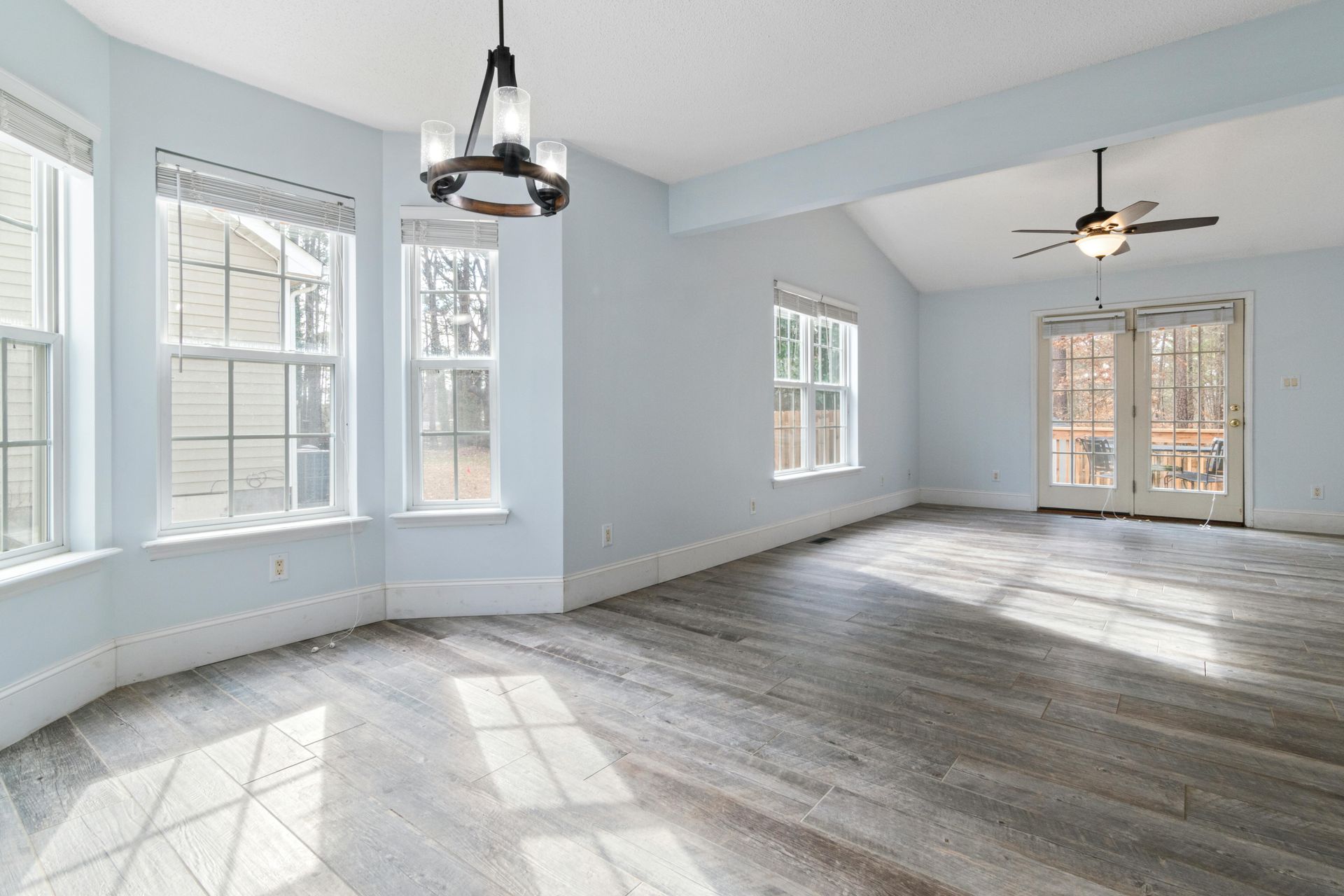 A bright, spacious room with light blue walls, gray wood-look flooring, large windows, a chandelier, and a ceiling fan.