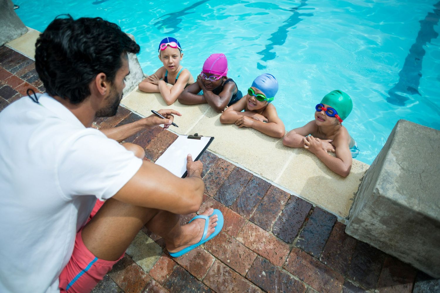 A man is sitting on the edge of a swimming pool talking to a group of children.