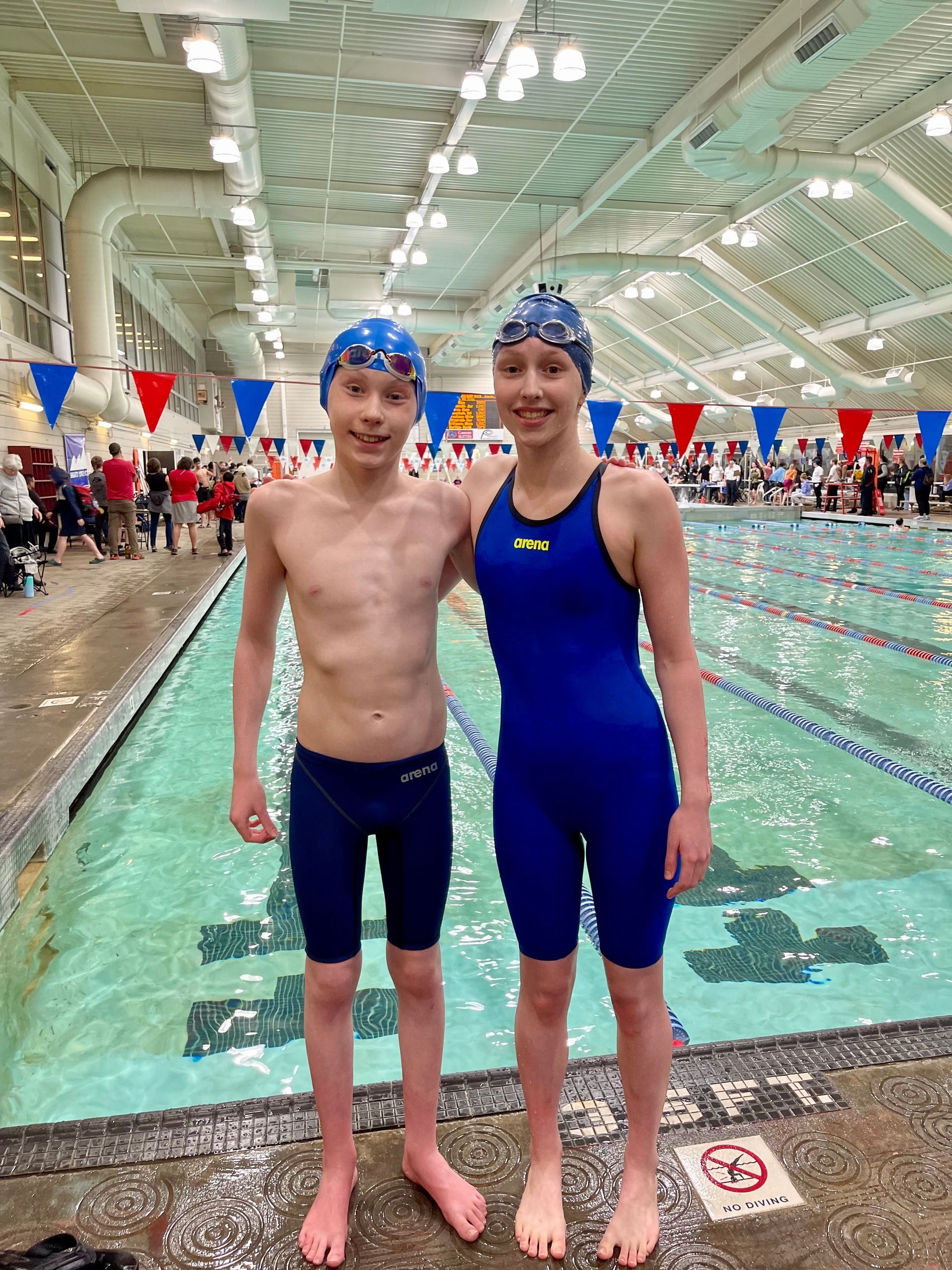 a boy and a girl are standing next to each other in front of a pool wearing arena swimsuits