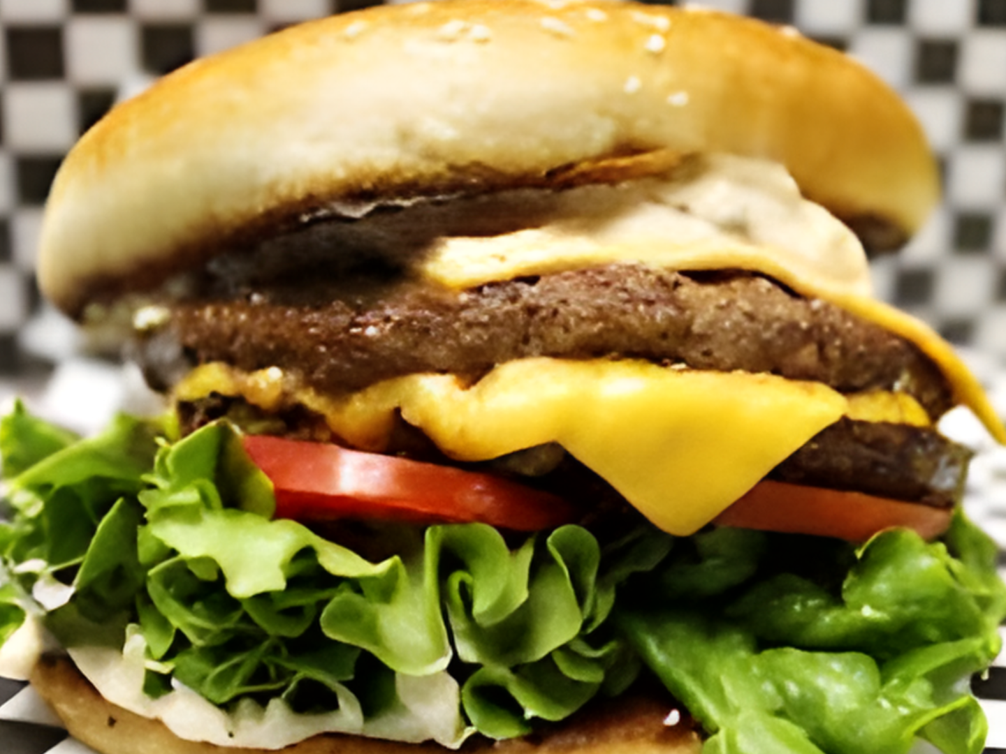 A hamburger with lettuce , tomatoes and cheese on a checkered table cloth.