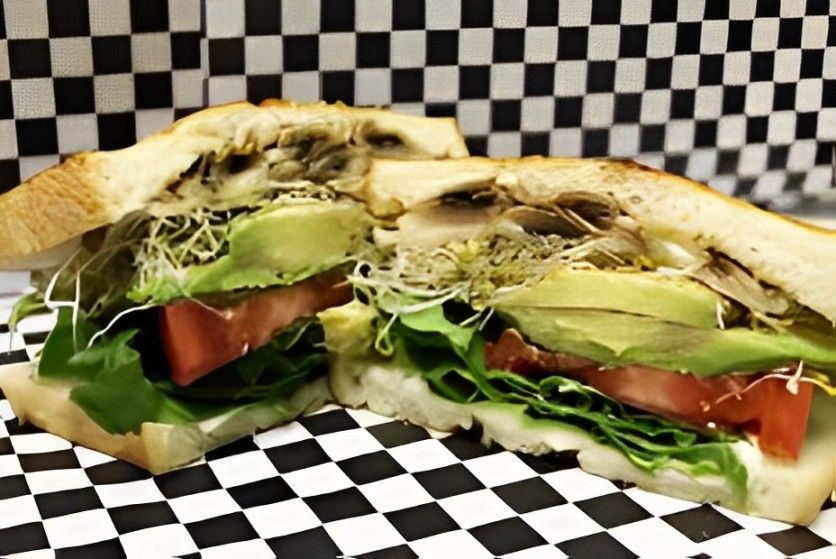 A sandwich with avocado , tomatoes , lettuce and mushrooms is cut in half on a checkered paper.