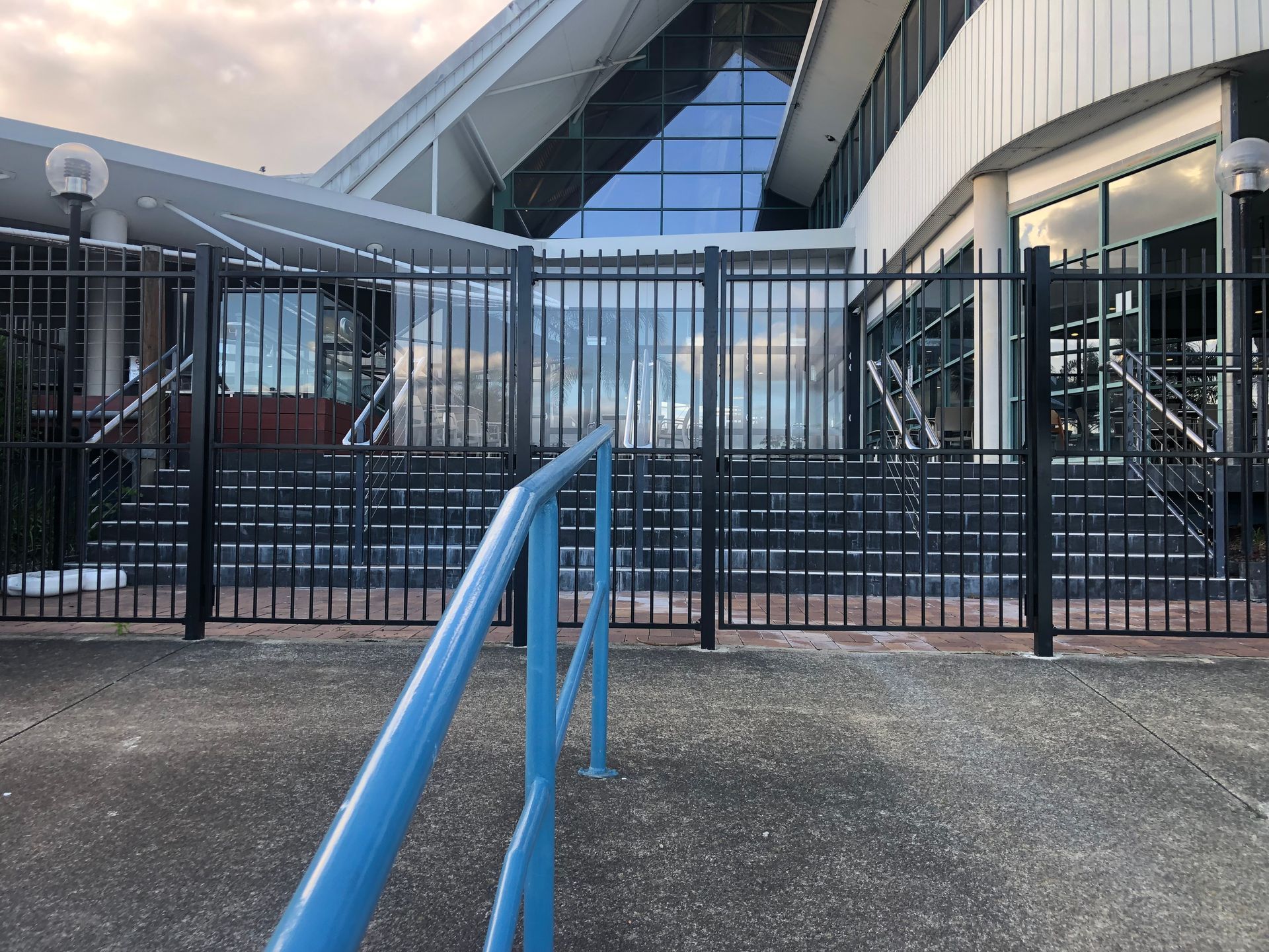 Exterior view of a building with a dark metal fence, steps, and blue handrail. The building has a triangular glass facade — Fencer Steve In Temagog, NSW