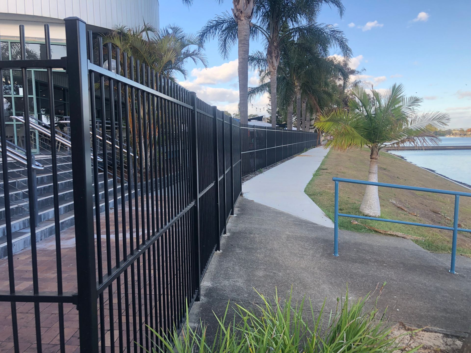 Black metal fence alongside a concrete pathway next to water, with palm trees in background — Fencer Steve In Temagog, NSW