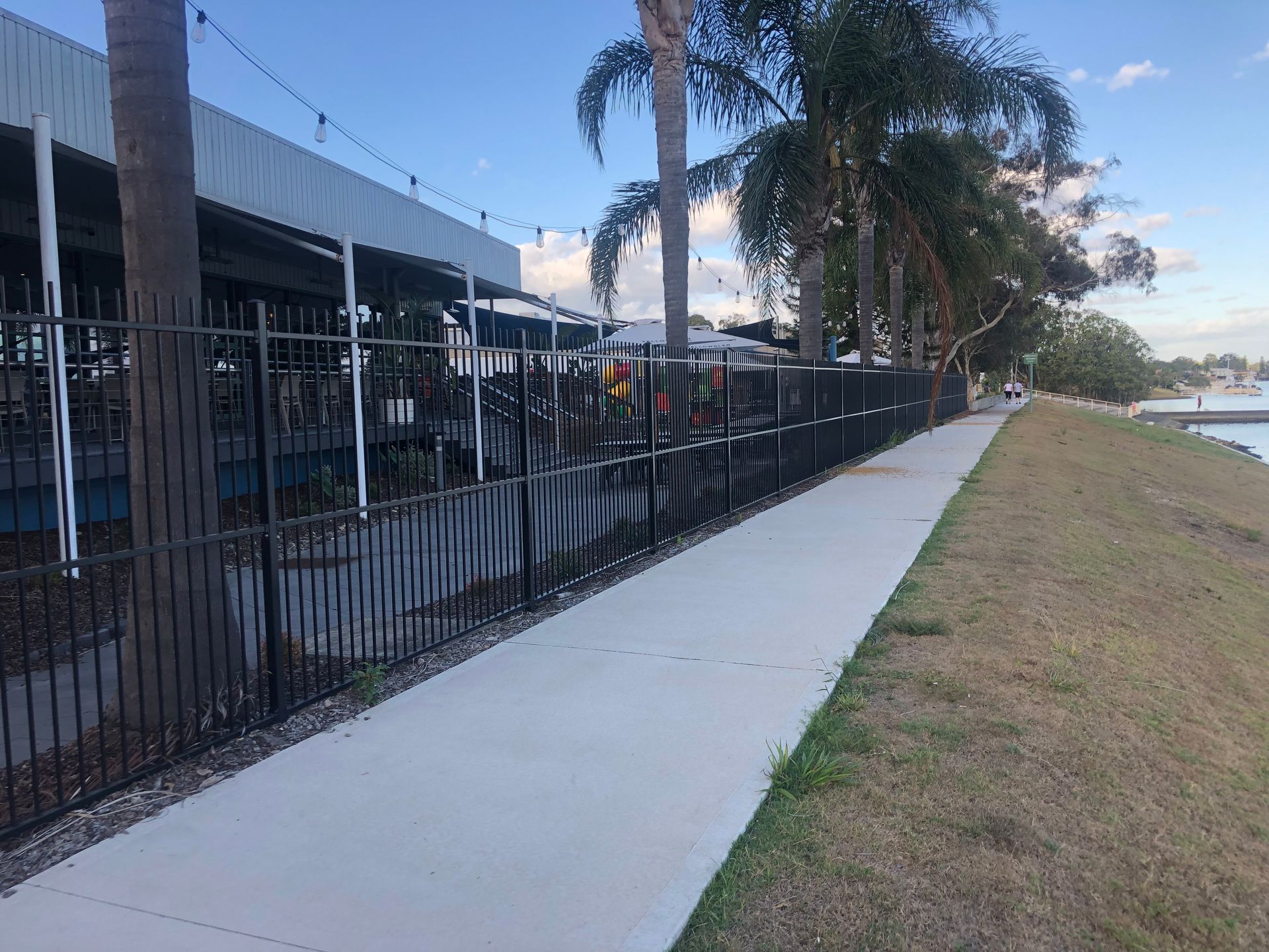 Black metal fence along a paved sidewalk next to a building and grassy embankment under a blue sky — Fencer Steve In Temagog, NSW