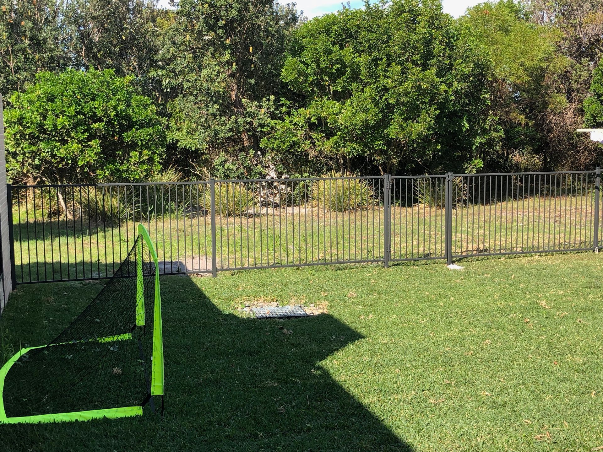 Green soccer goal on a grassy lawn, black fence and trees in the background. Sunny day.