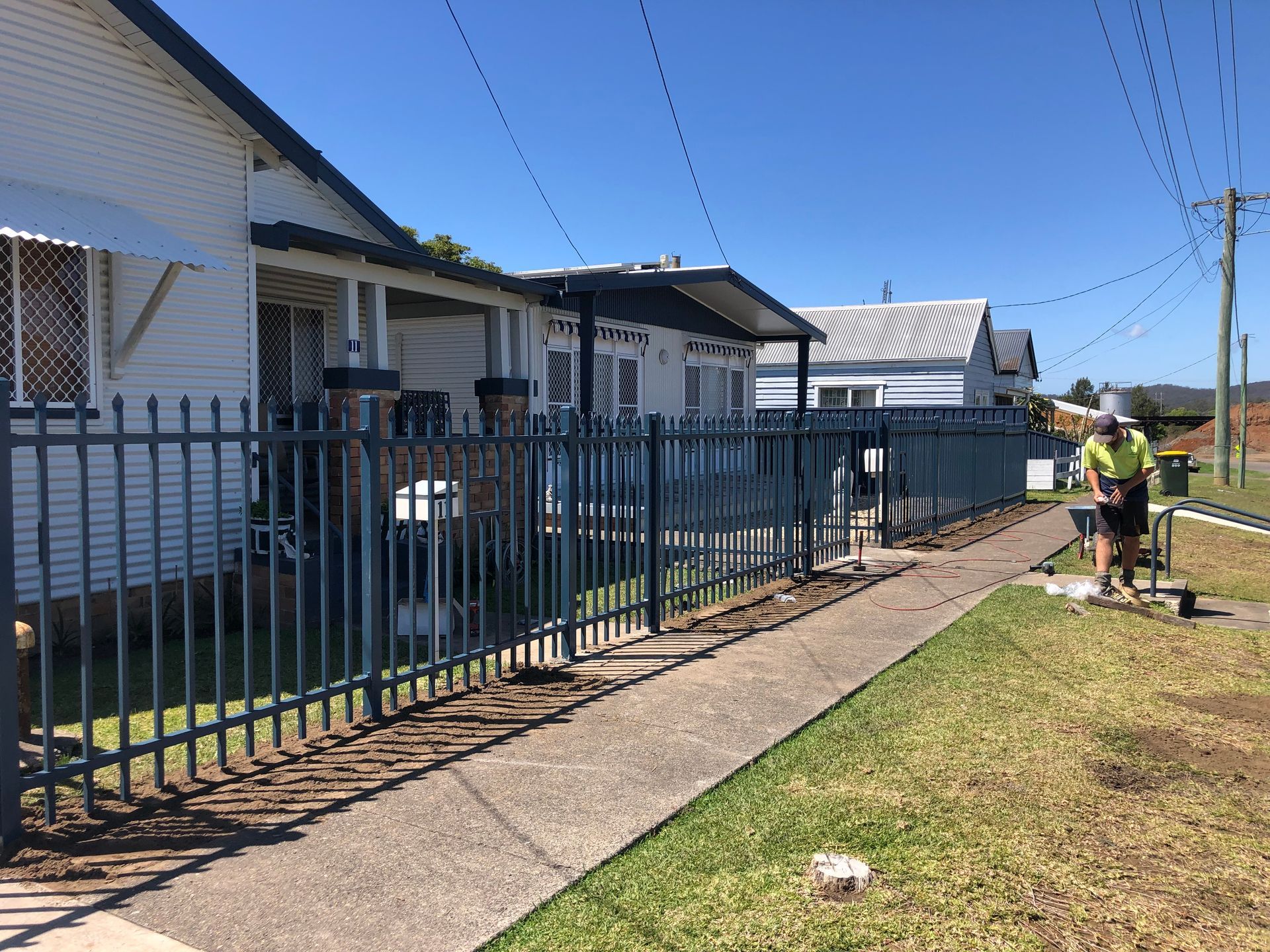 A blue metal fence bordering a sidewalk in front of several houses. A person works near the end — Fencer Steve In Temagog, NSW