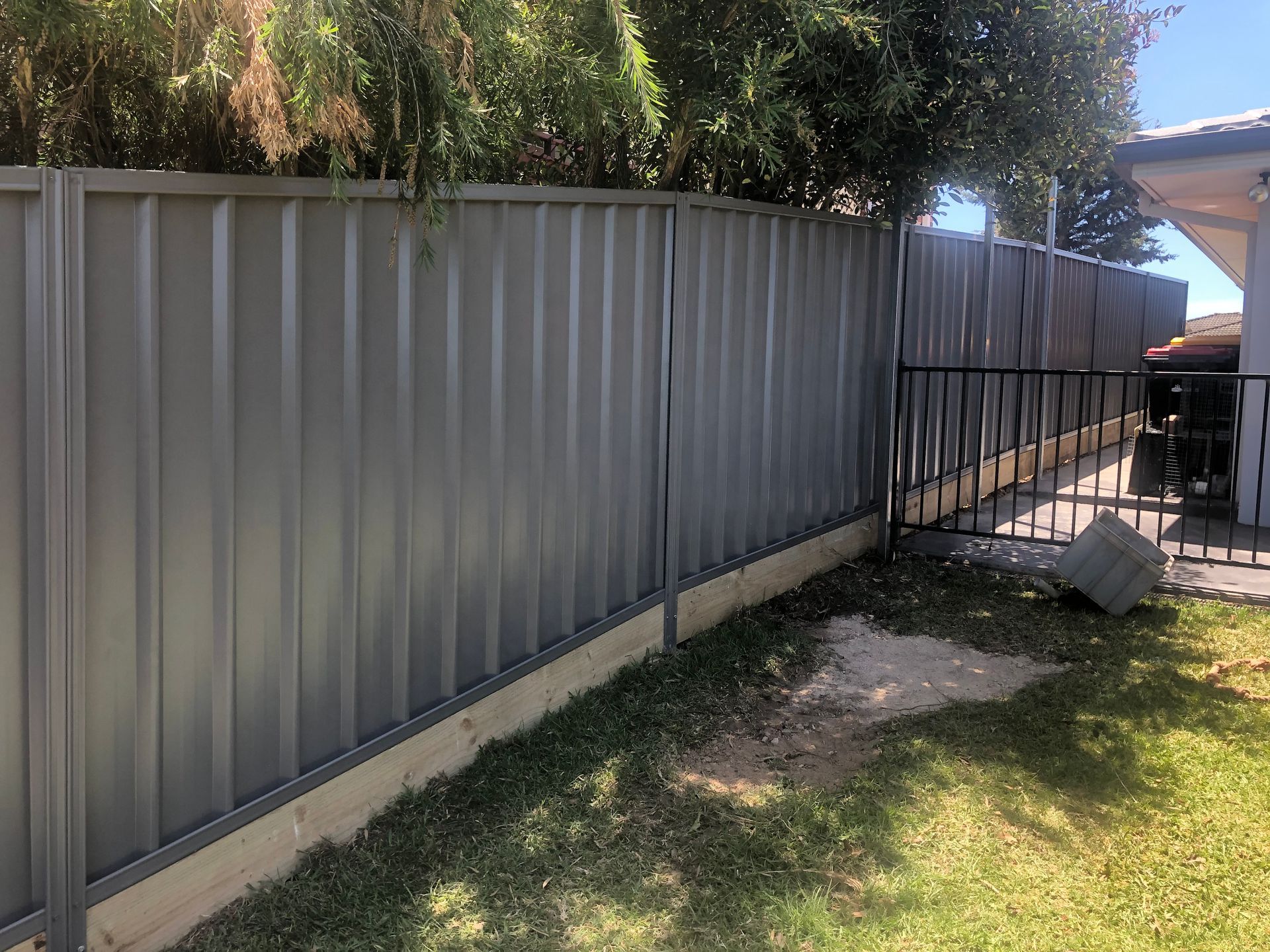 A gray corrugated metal fence with a wooden base in a backyard, with a black metal gate — Fencer Steve In Temagog, NSW