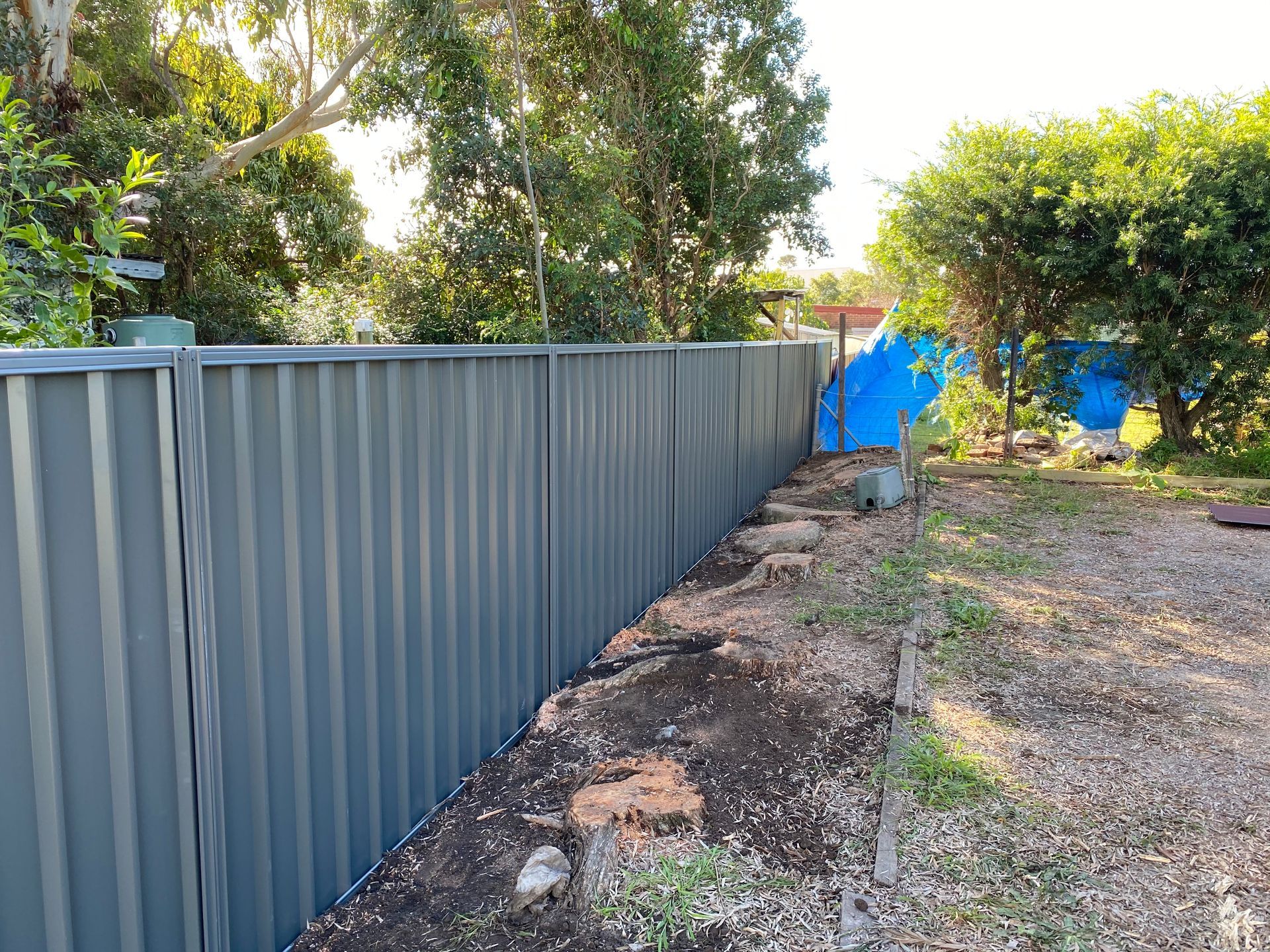 A gray corrugated metal fence in a backyard, with a dirt and brick border. Trees in the background — Fencer Steve In Temagog, NSW
