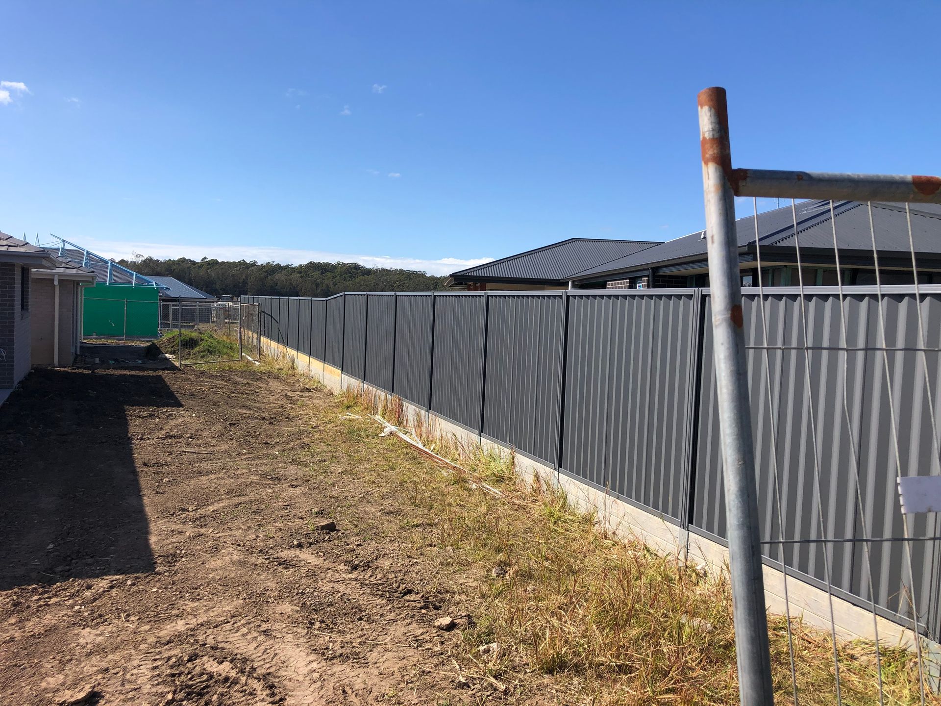 Dirt path with a dark gray corrugated fence, blue sky, and houses — Fencer Steve In Temagog, NSW