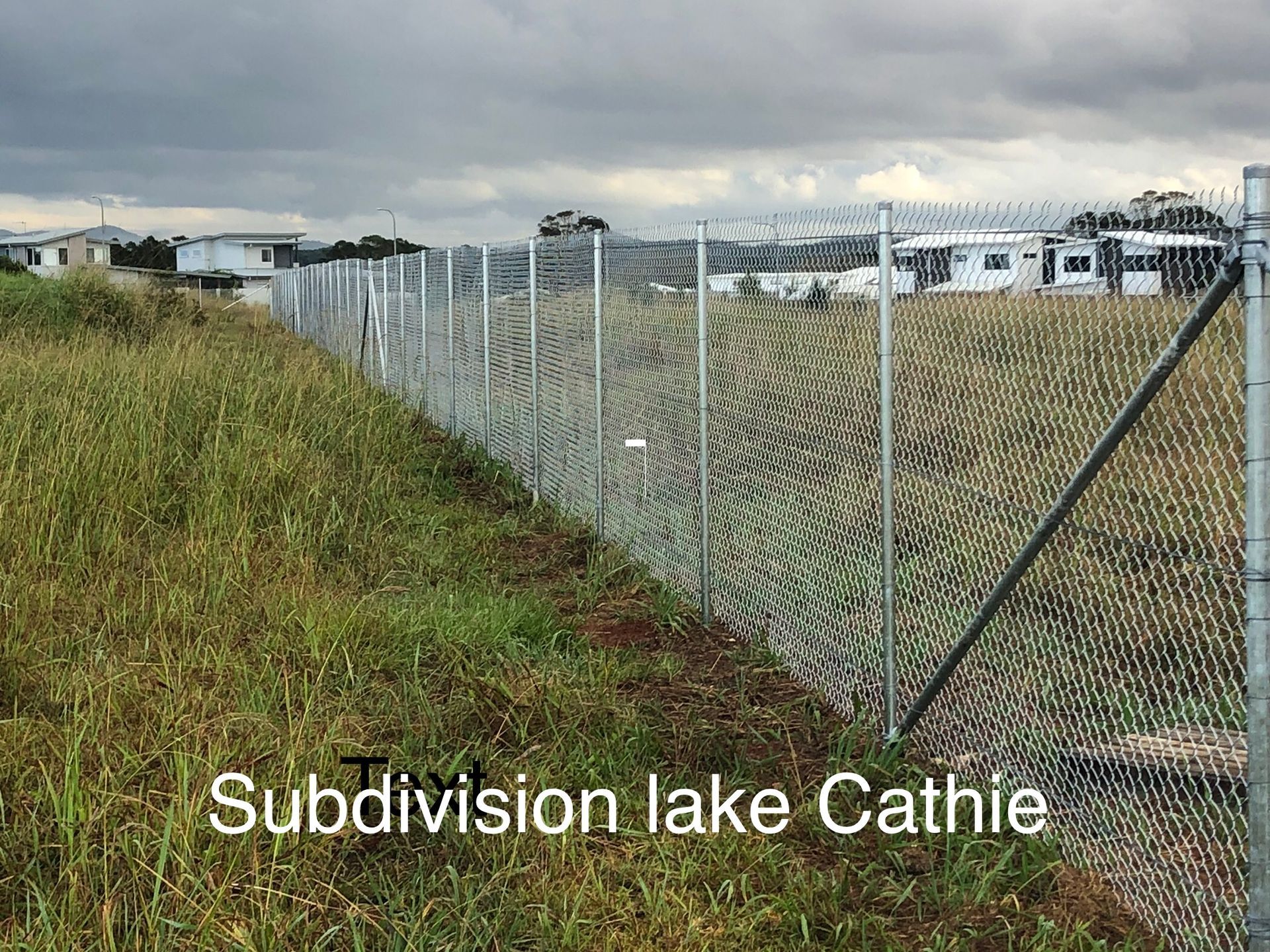 Chain-link fence along Subdivision lake Cathie, with long grass in the foreground and houses in the distance. Cloudy sky — Fencer Steve In Temagog, NSW