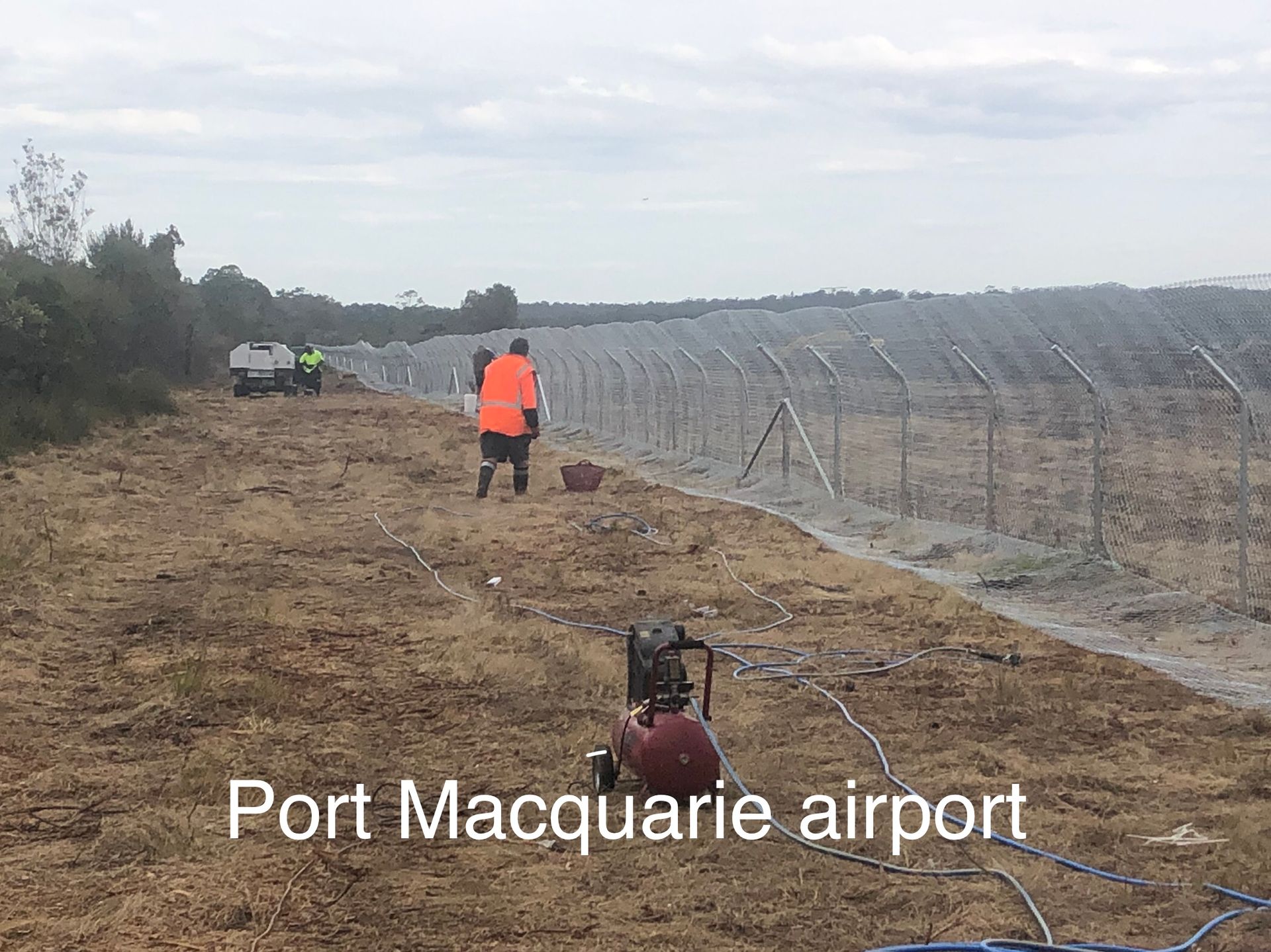 Workers near Port Macquarie airport, preparing ground with equipment and installing fencing, under a cloudy sky — Fencer Steve In Temagog, NSW