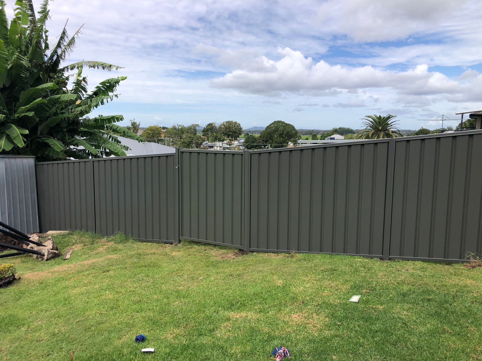 Gray corrugated metal fence in a yard with green grass, trees, and a cloudy sky — Fencer Steve In Temagog, NSW