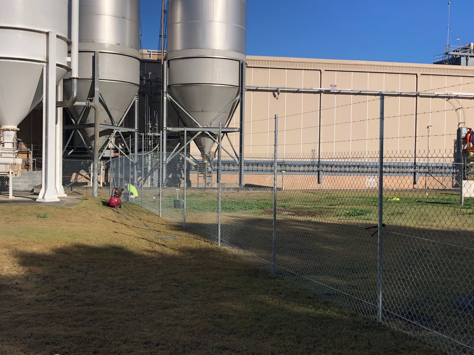 Chain-link fence being installed near industrial silos and a building, two workers nearby on a sunny day — Fencer Steve In Temagog, NSW
