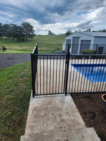 Black fence with gate surrounds a pool, concrete path, and grassy landscape under a cloudy sky — Fencer Steve In Temagog, NSW