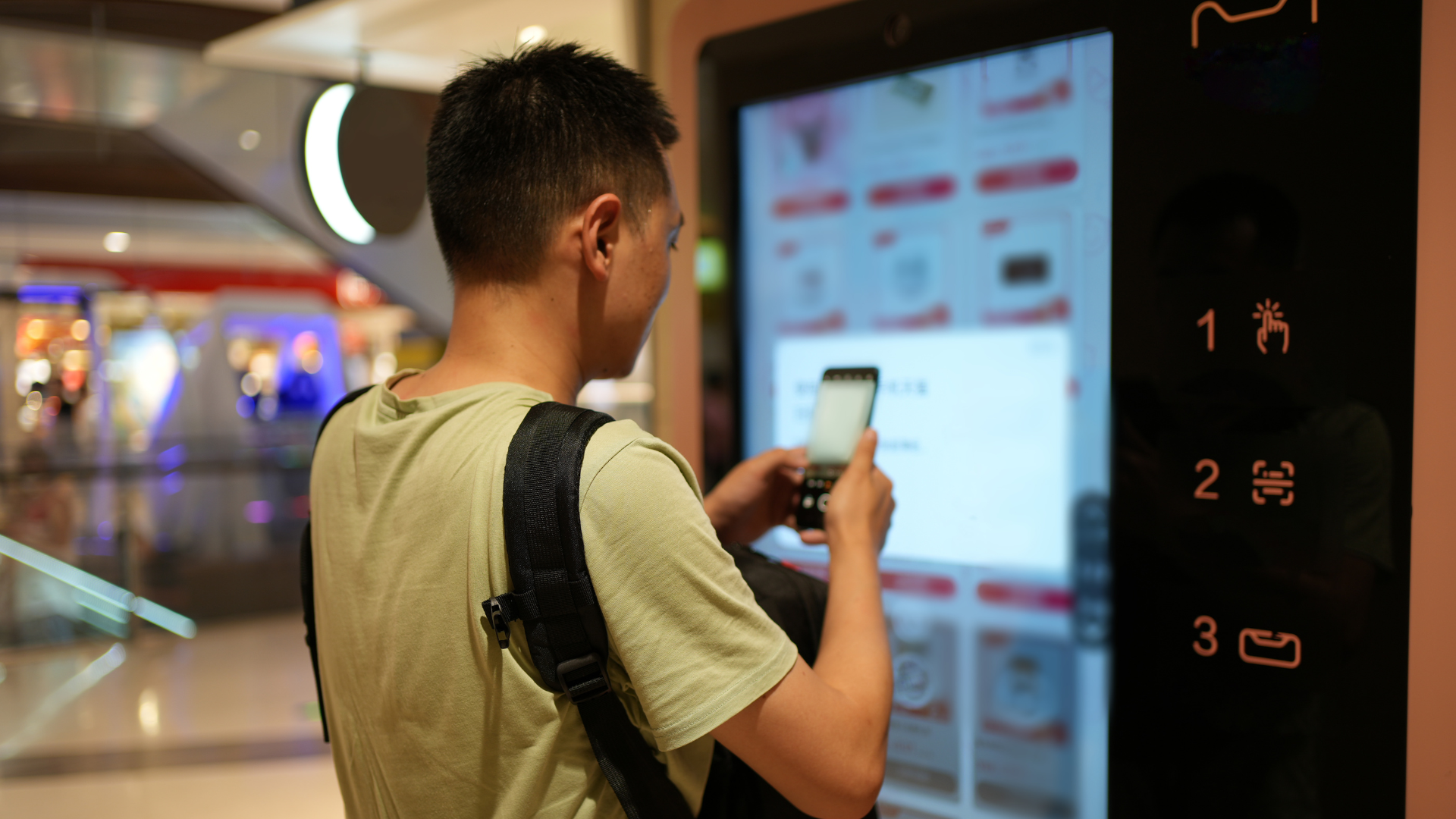 Person using a smartphone at a touchscreen kiosk in a shopping mall corridor