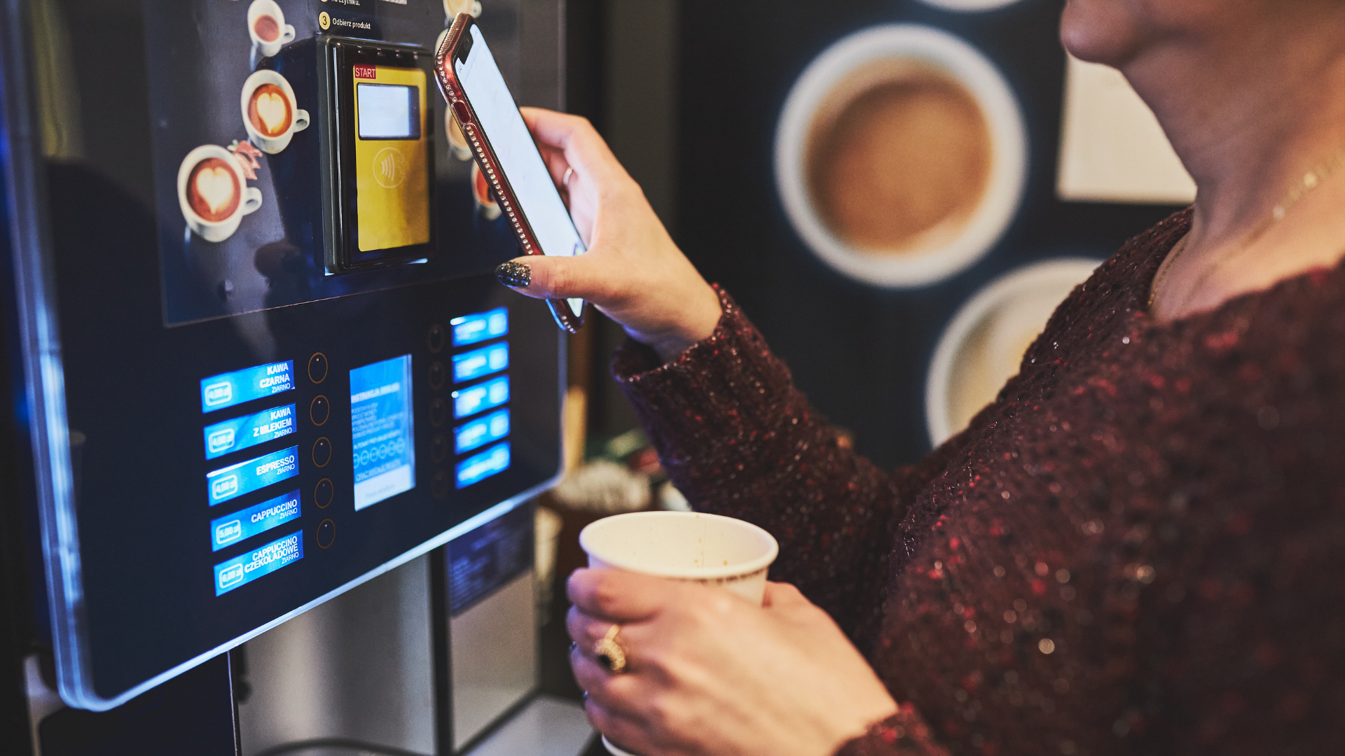 Person using a touchscreen coffee machine while holding a cup in a café