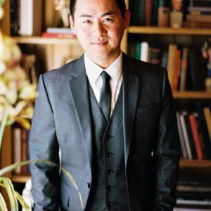 A man in a suit and tie is standing in front of a bookshelf.
