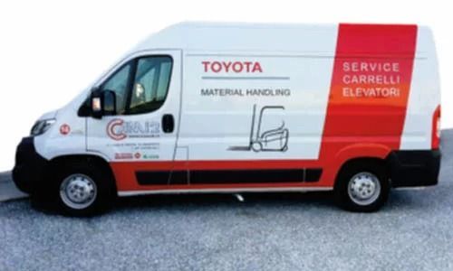 Vista laterale di un furgone bianco con il marchio Toyota Material Handling e la grafica di un carrello elevatore, parcheggiato sulla ghiaia.