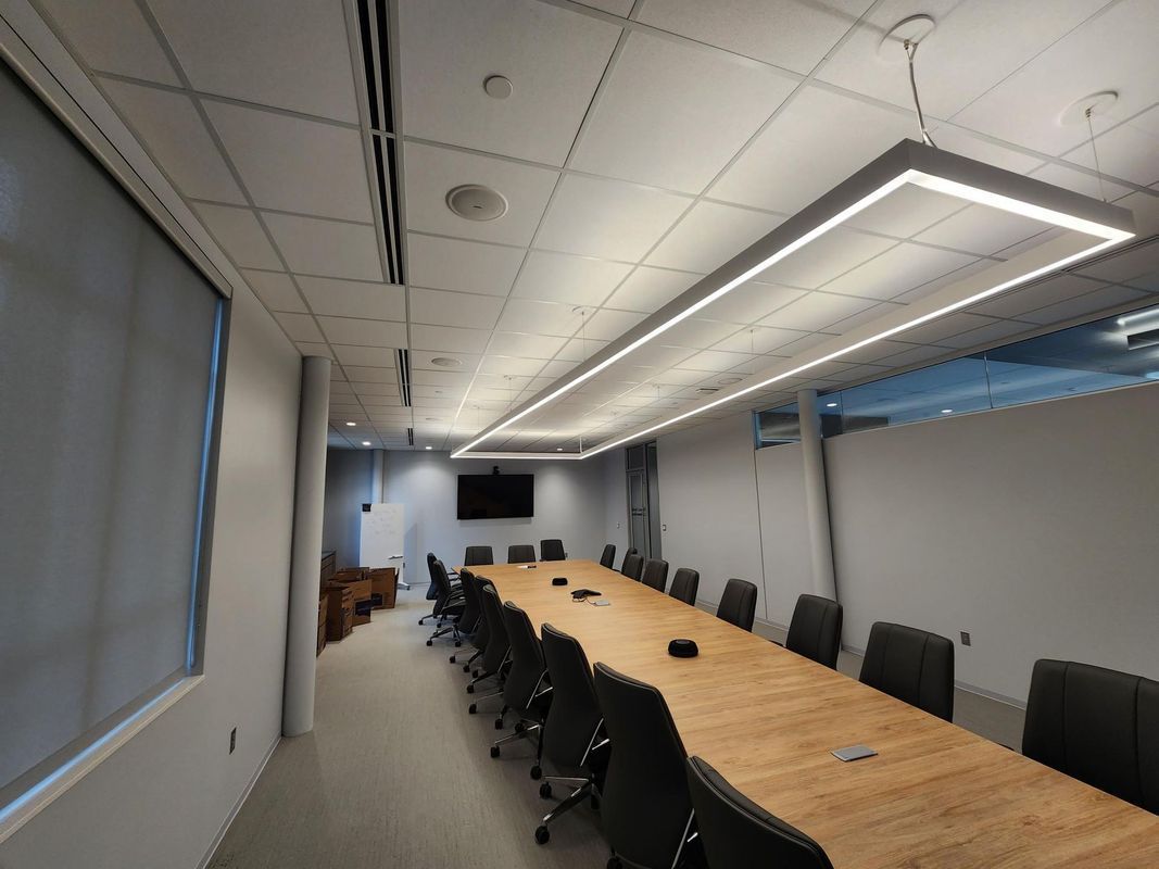 Conference room with long wooden table, black chairs, and overhead lighting. Gray walls, window with shade, and a large TV.