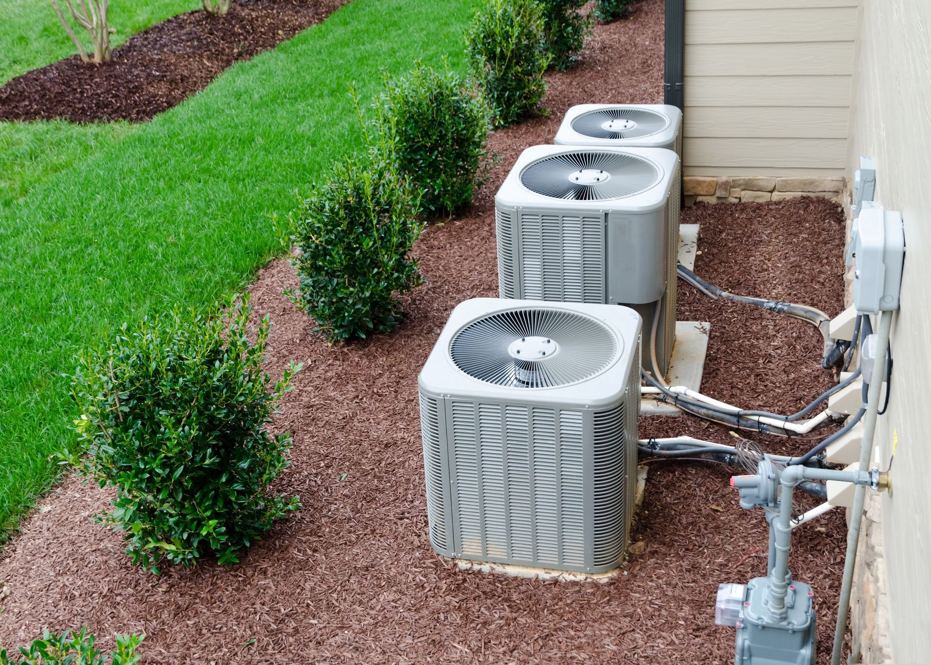 Three air conditioning units outside a building on brown mulch next to green shrubs and grass.