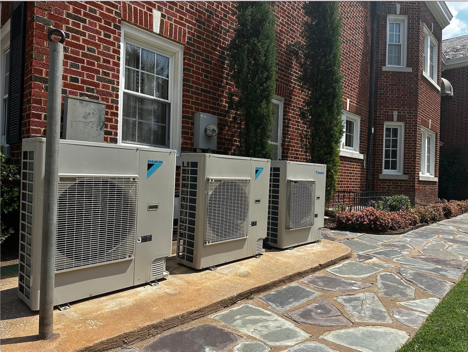 Three Daikin HVAC units lined up against a red brick building with stone walkway.