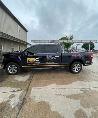 Dark blue RBC roofing truck parked next to a building with a ladder on top.