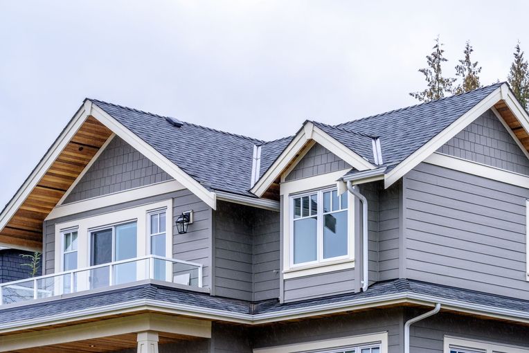 Gray house with white trim, glass balcony, blue roof against a cloudy sky.