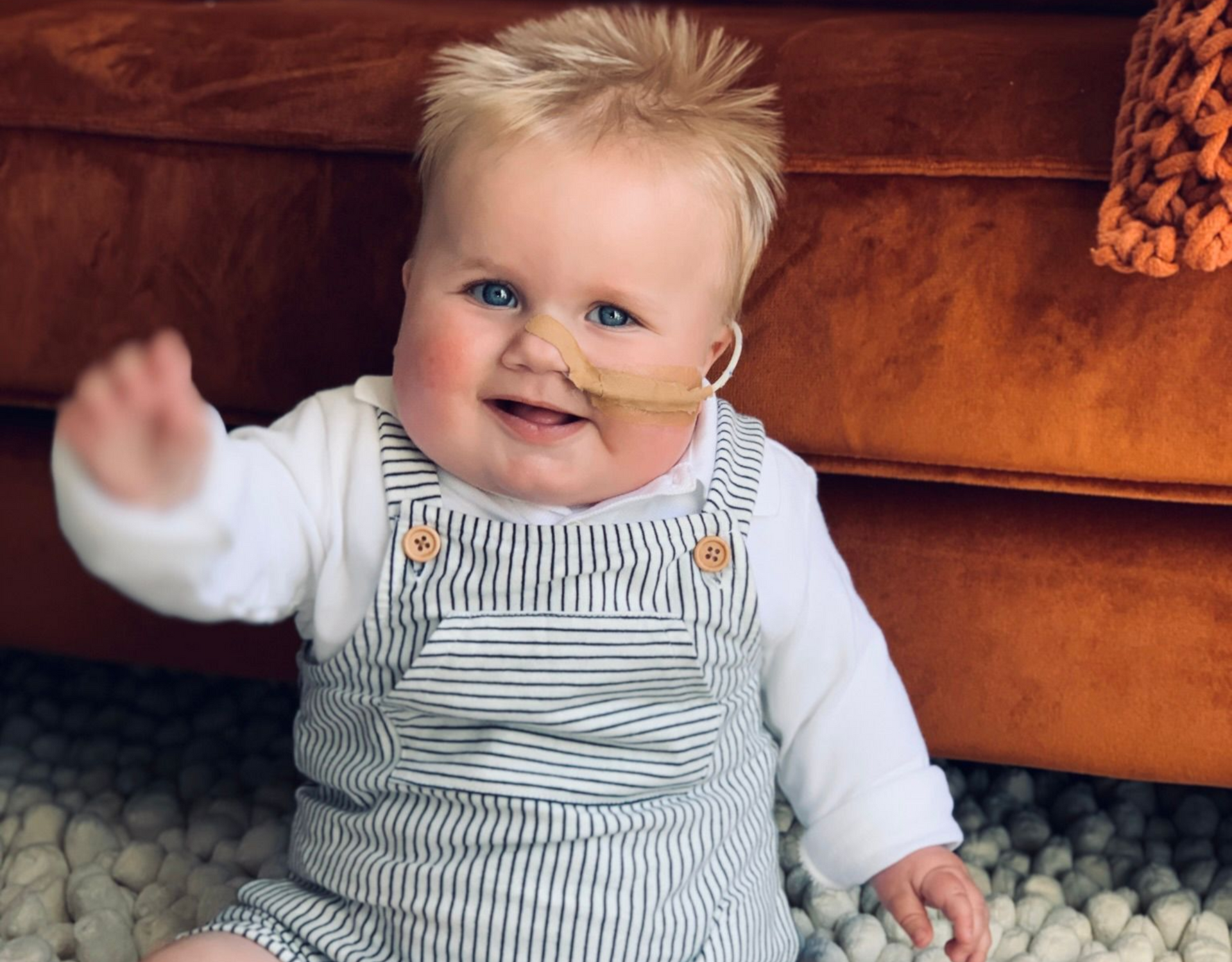 Een lachende baby met blond haar, een gestreepte overall aan en een voedingssonde, zwaait.
