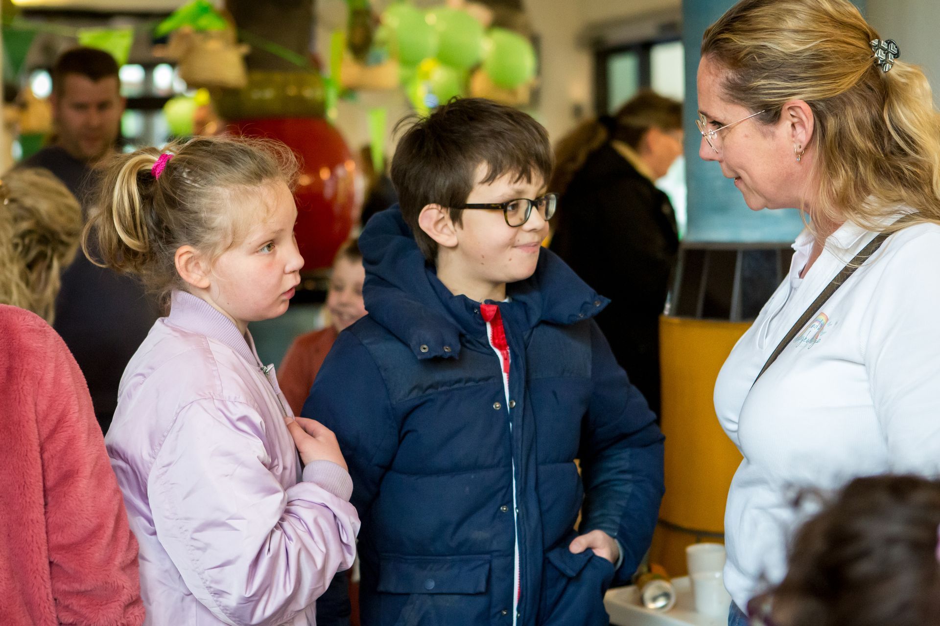 Twee kinderen praten met een vrouw binnenshuis; een jongen in een blauwe jas, een meisje