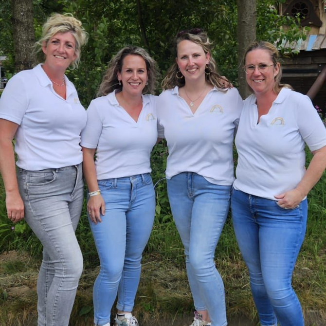 Vier vrouwen in witte shirts en spijkerbroeken poseren samen in de buitenlucht.