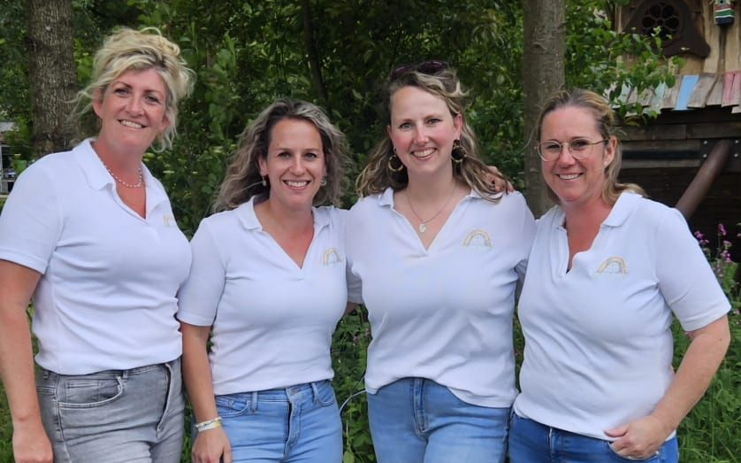 Vier vrouwen in witte shirts en spijkerbroeken poseren lachend in de buitenlucht.