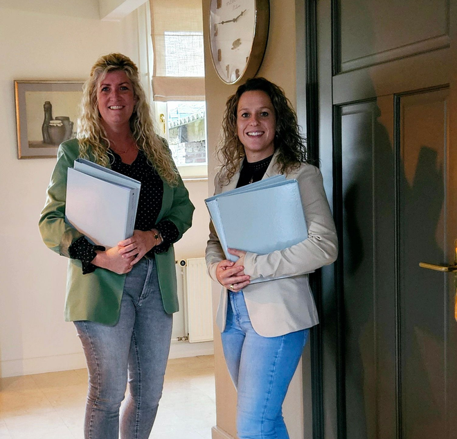 Twee vrouwen in zakelijke kleding, met mappen in hun handen, staan ​​glimlachend in een deuropening van een kantoor.